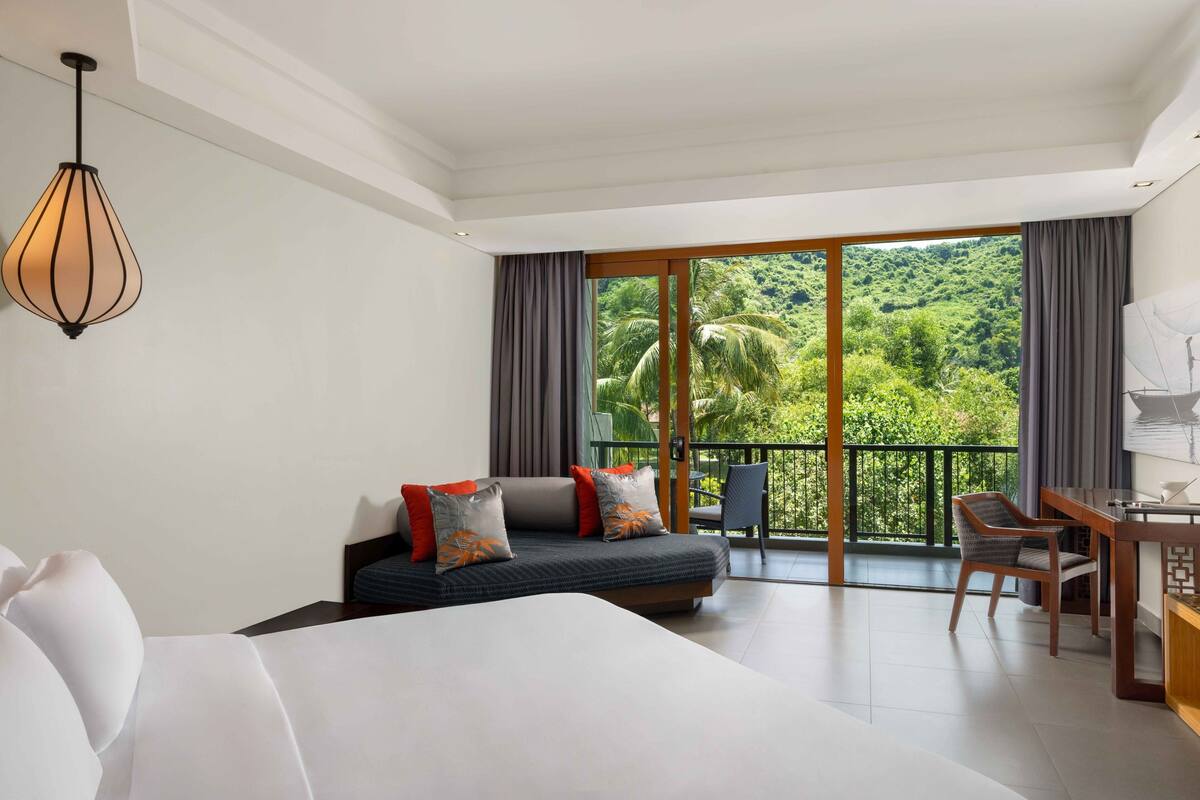 A hotel room with a bed, a sofa, a desk, and a chair. There is a large window with a view of greenery outside.