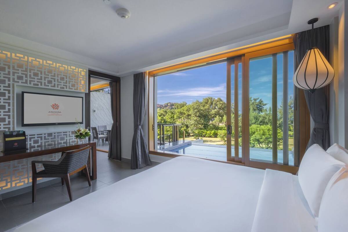 A hotel room with a large bed, a desk with a chair, a TV mounted on the wall, and a balcony with a pool.