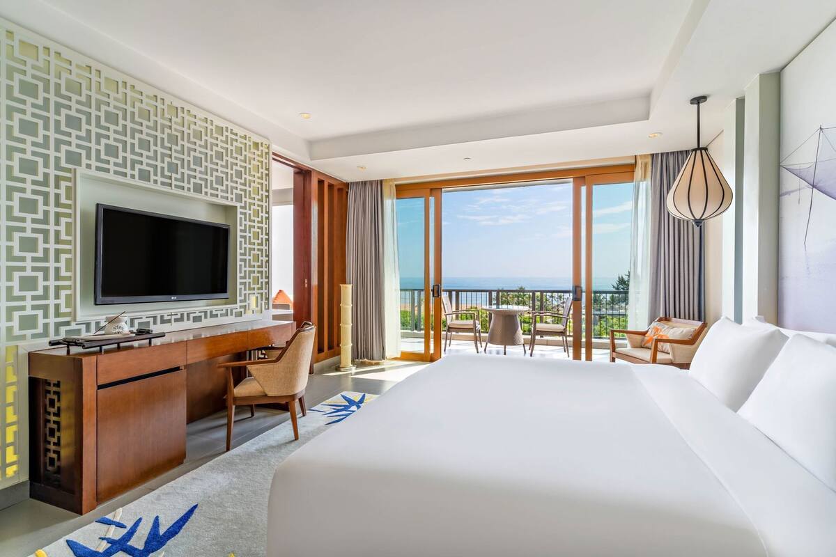 A hotel room with a large bed, a desk with a TV, a balcony with a table and chairs, and a view of the ocean.