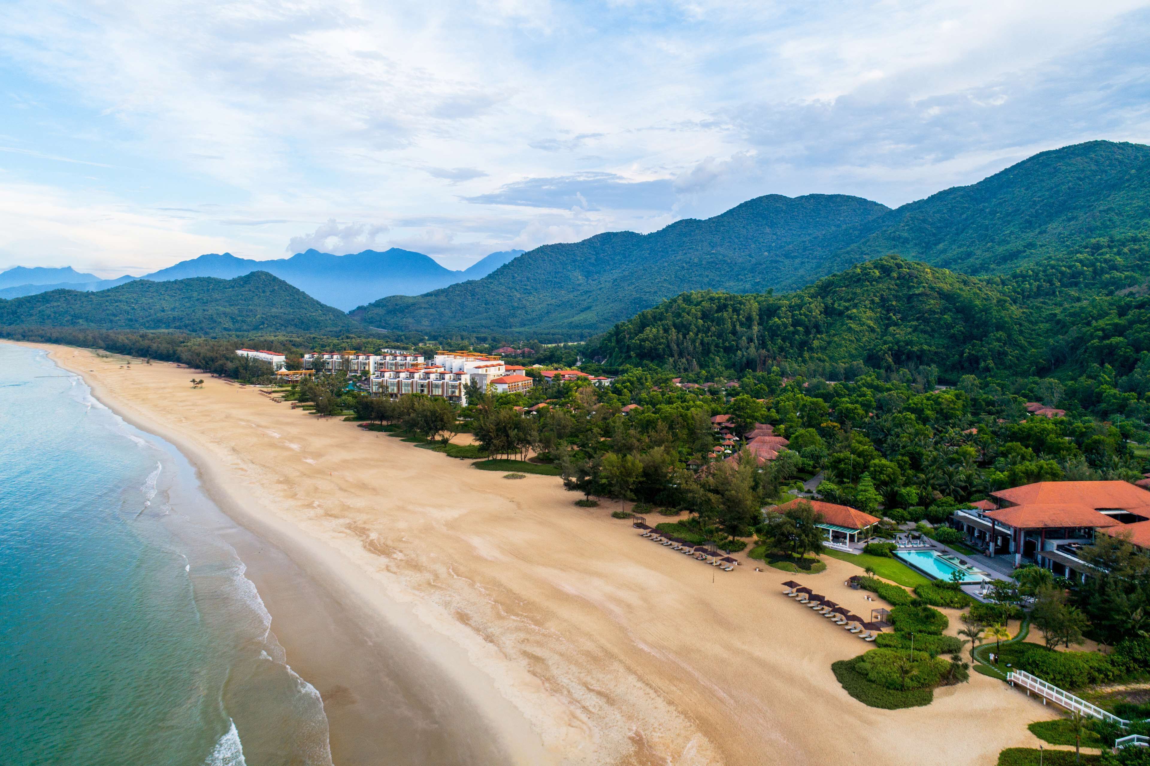 Un complejo turístico frente al mar, con playa, montañas al fondo y varias edificaciones con tejados rojos.