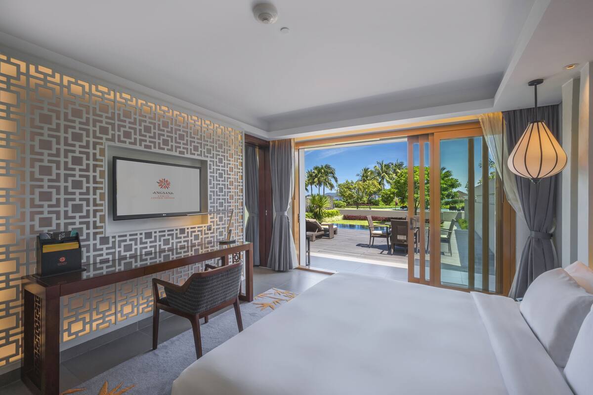 A modern hotel room with a large bed, a desk with a chair, a TV mounted on the wall, and a view of the outdoor pool area.