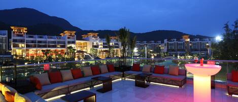 Un área de lounge en la azotea con vista a la piscina, mobiliario exterior y una zona de bar bien iluminada.
