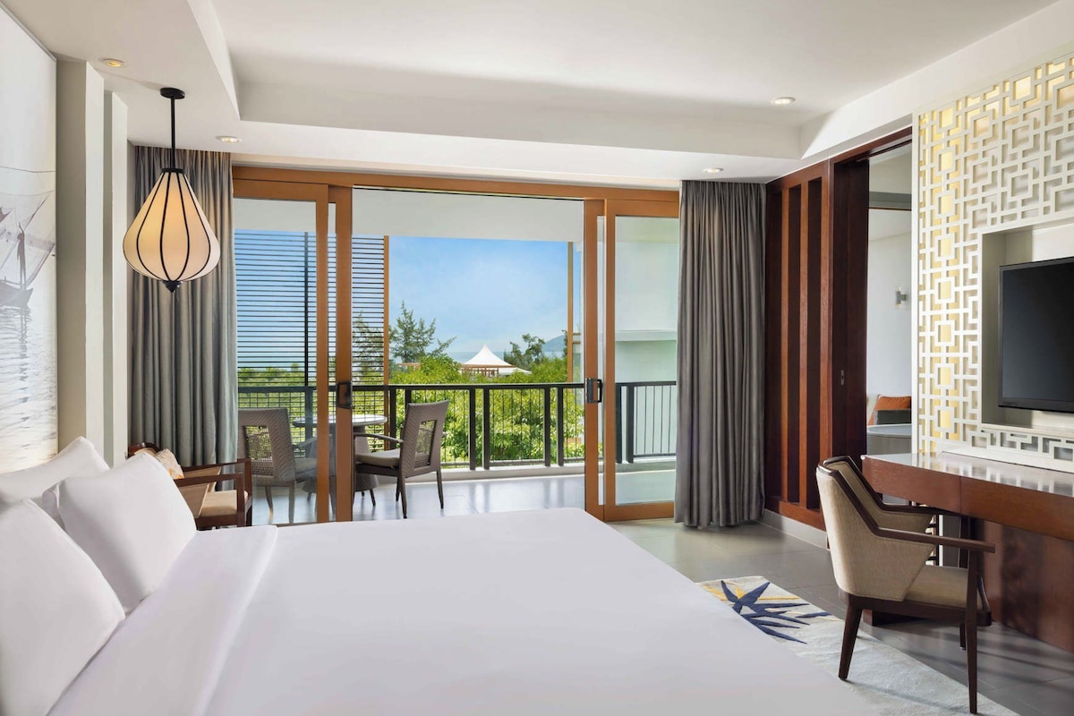 A hotel room with a large bed, a desk, a TV, and a balcony with a view.
