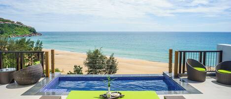 A pool area with a view of the beach and ocean, surrounded by wicker furniture and a tiled deck.