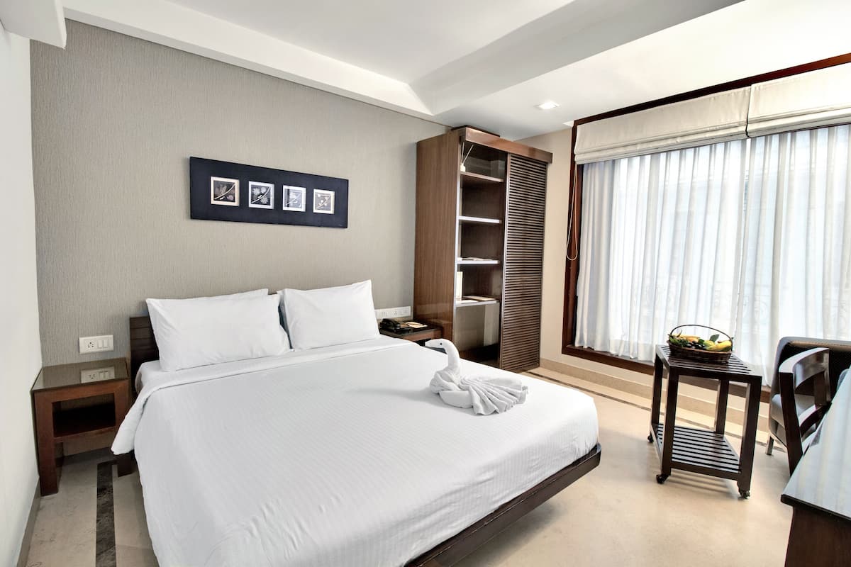 A hotel room with a large bed, a wooden wardrobe, a small table with a fruit basket, and a window with curtains.