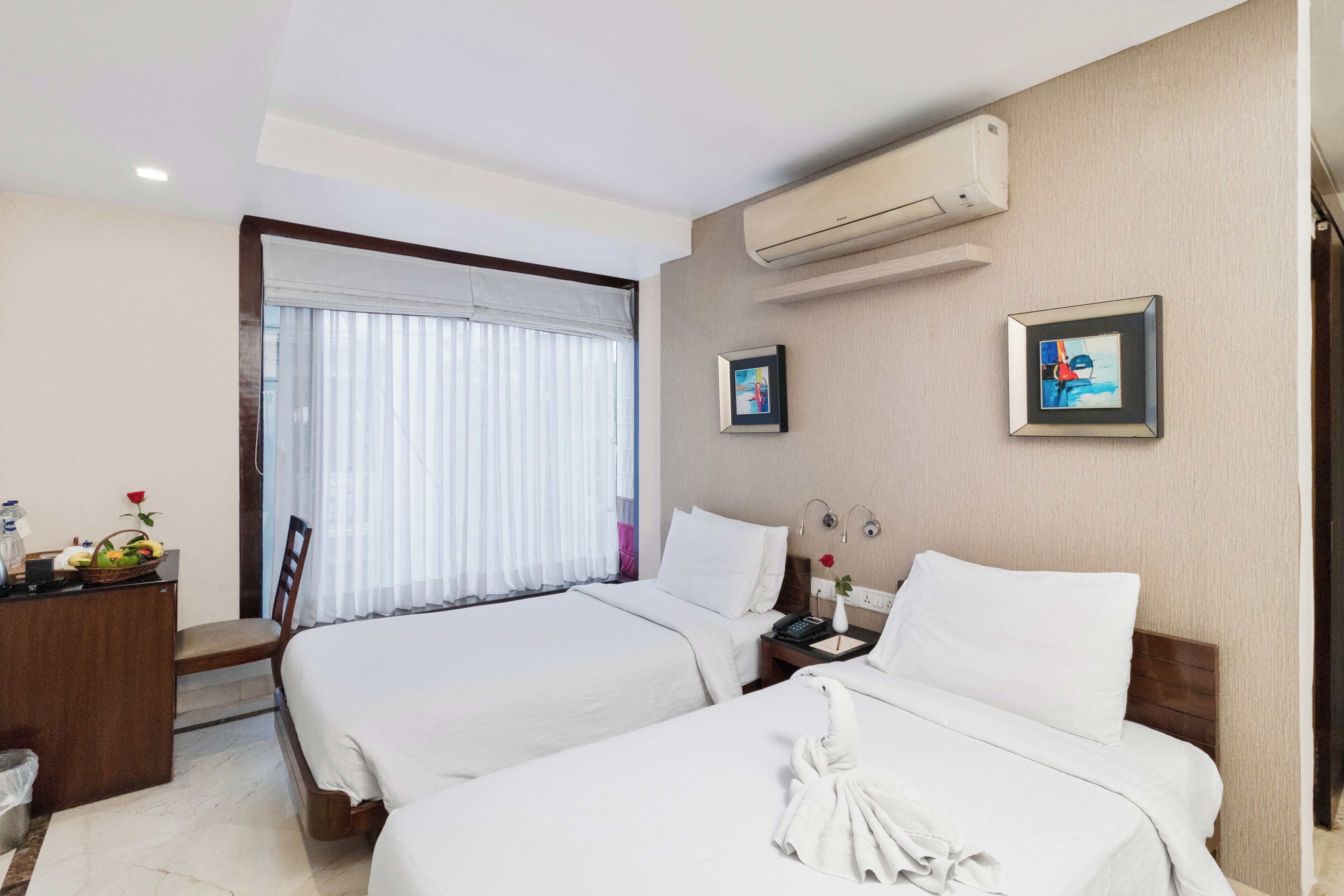 Habitación de hotel con dos camas, un escritorio, una silla, una ventana con persianas, un equipo de aire acondicionado y dos cuadros en la pared.