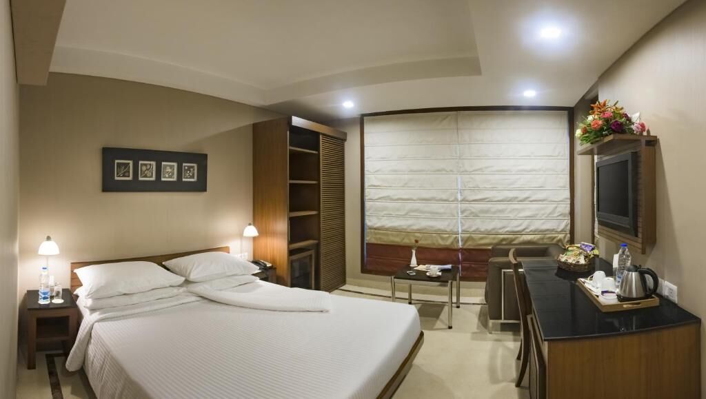 A hotel room with a bed, bedside table, lamp, desk, chair, and a large window with curtains.