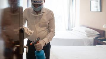 Une personne se trouve dans une chambre d’hôtel, tenant un vaporisateur et un chiffon de nettoyage, et se tient près d’une porte.