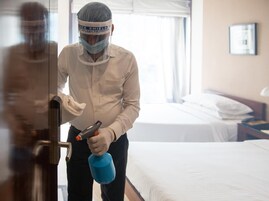 A person in a hotel room holding a spray bottle and a cleaning cloth, standing near a door.