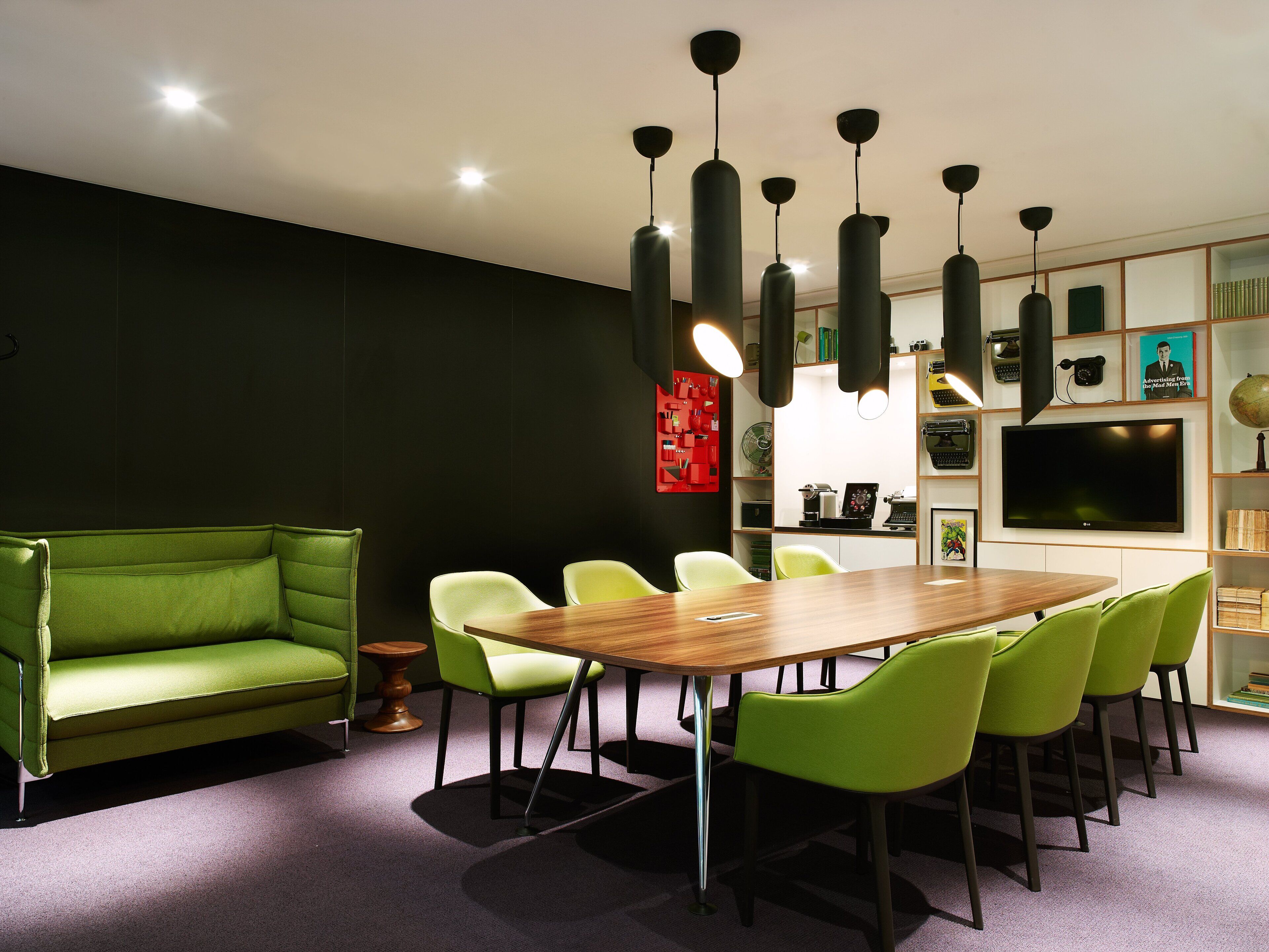 A modern meeting room with a wooden table, green chairs, a green sofa, and pendant lighting.