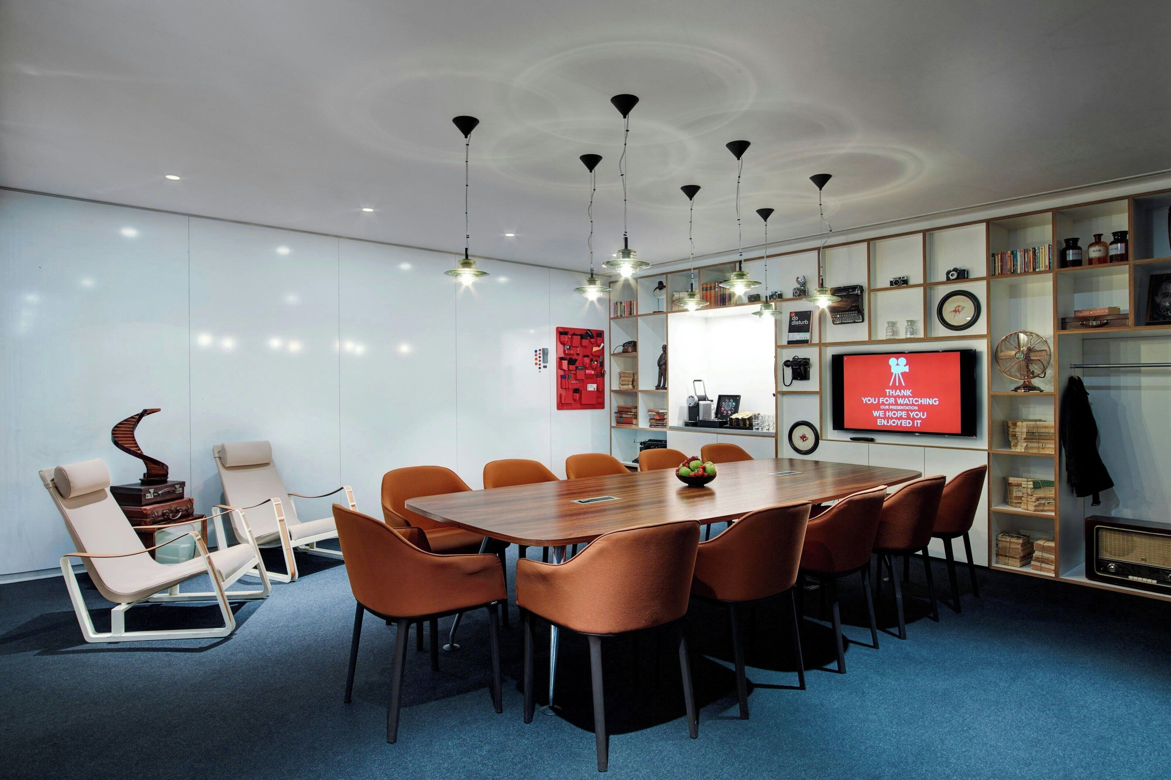 A conference room with a long table, orange chairs, a television screen, and a bookshelf with various items.