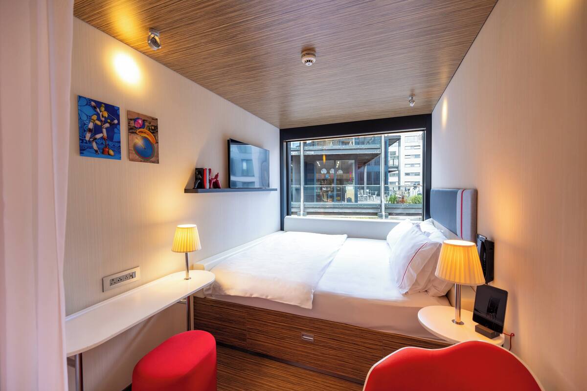 A modern hotel room with a bed, bedside table, lamp, and red chairs.