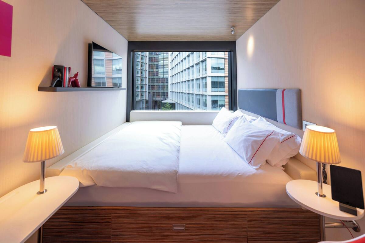 A modern hotel room with a large bed, bedside tables, and a window with a city view.