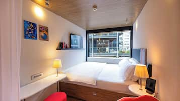 A modern hotel room with a bed, bedside tables, a desk, and a window with a city view.