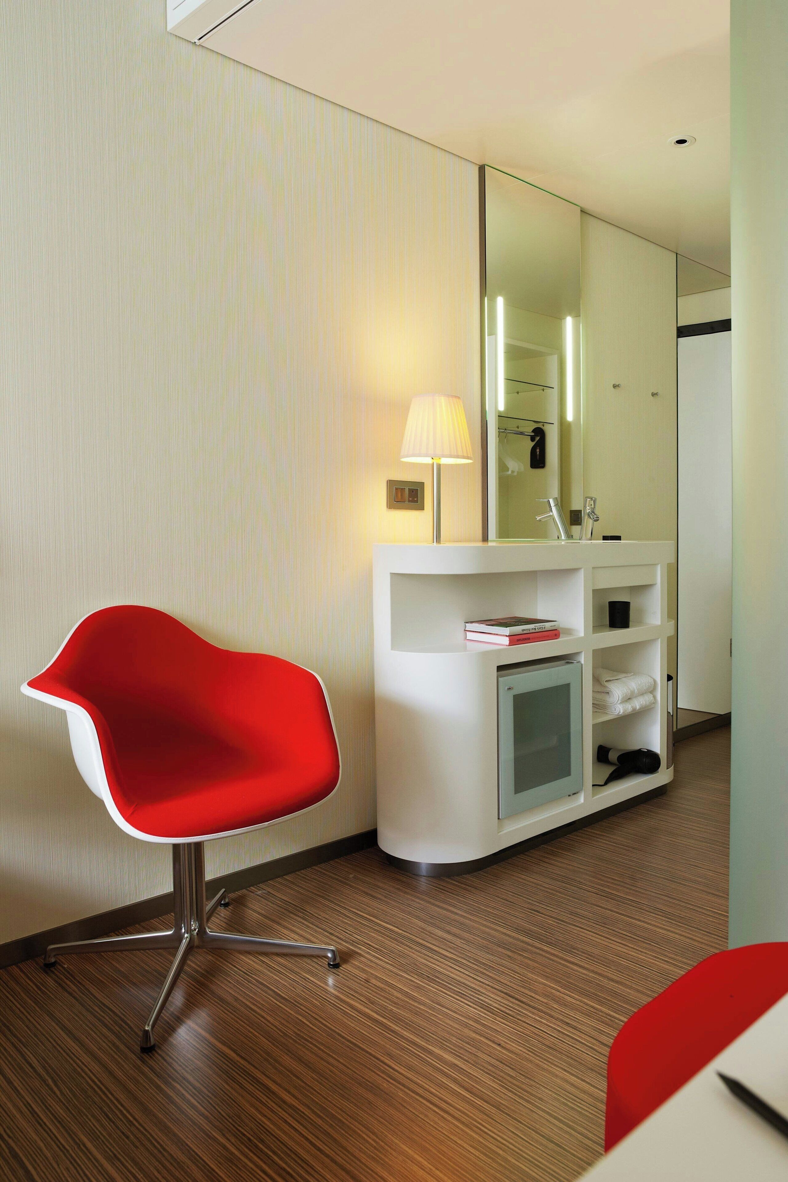 A modern hotel room with a red chair, a white vanity with a mirror, and a small refrigerator.