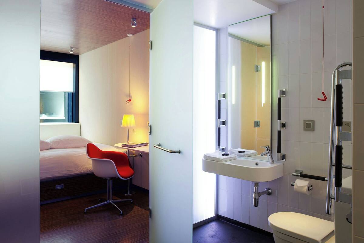 A modern hotel room with a bed, a red chair, a bedside table with a lamp, and a bathroom with a sink, mirror, and shower.