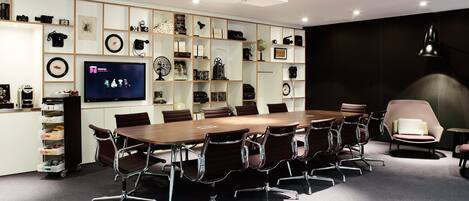 A modern meeting room with a large table, conference chairs, a television, and a bookshelf with various items.