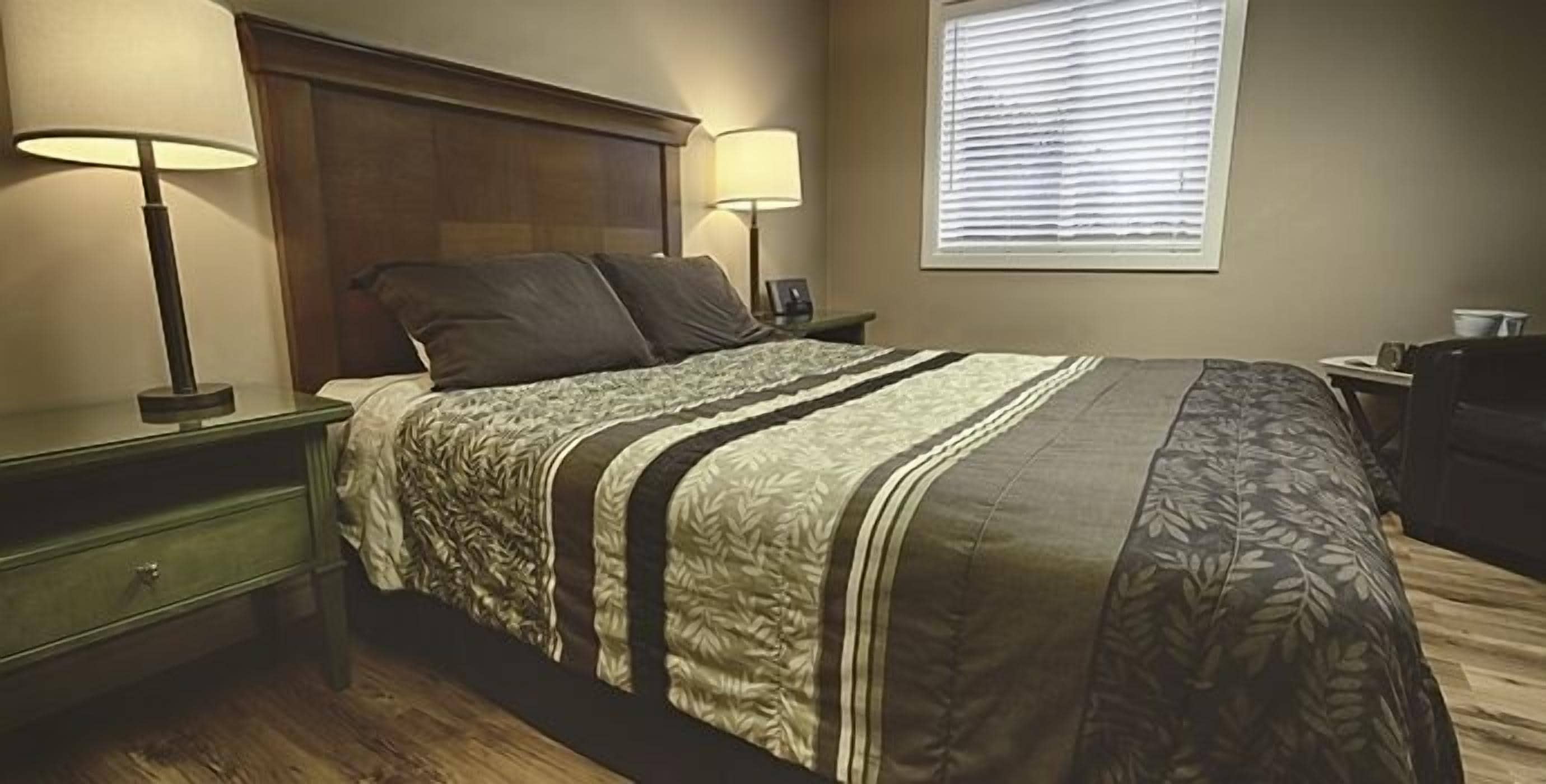 Una cama bien hecha con una colcha a rayas, dos lámparas de noche, un cabecero de madera y una ventana con persianas.