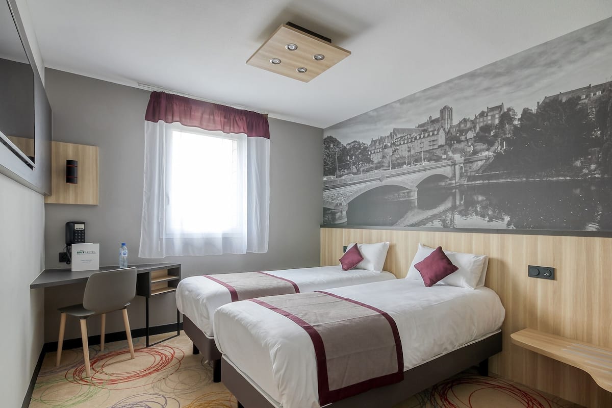 Habitación de hotel con dos camas, un escritorio, una silla, una ventana con cortina y un mural en la pared.