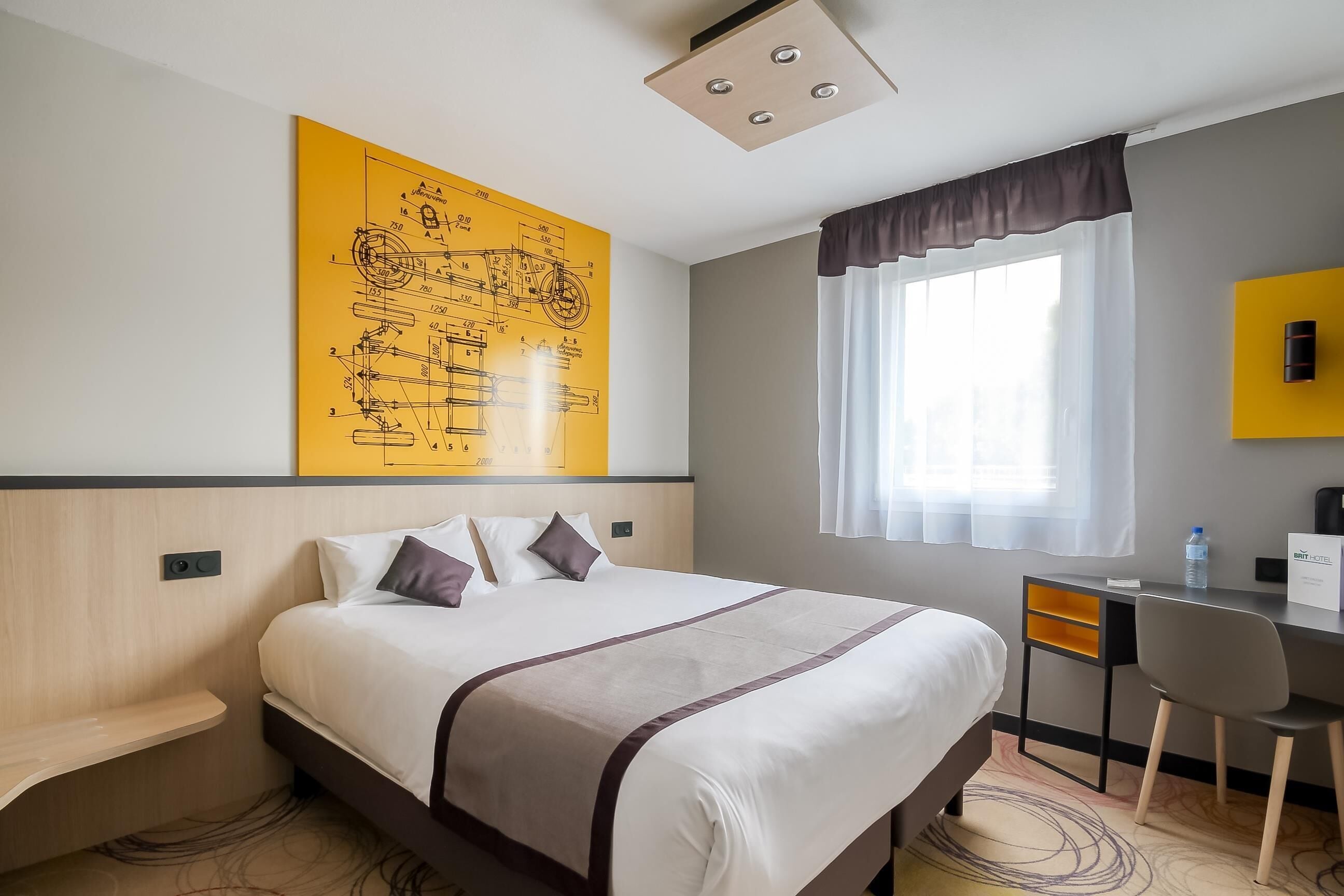 A hotel room with a bed, a desk, a chair, a large yellow wall art, a window with curtains, and a ceiling light.