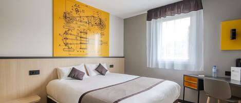 A hotel room with a bed, a desk, a chair, a large yellow wall art, a window with curtains, and a ceiling light.