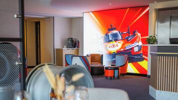 A modern hotel lobby with a reception desk, seating area, and a large digital screen displaying a racing theme.