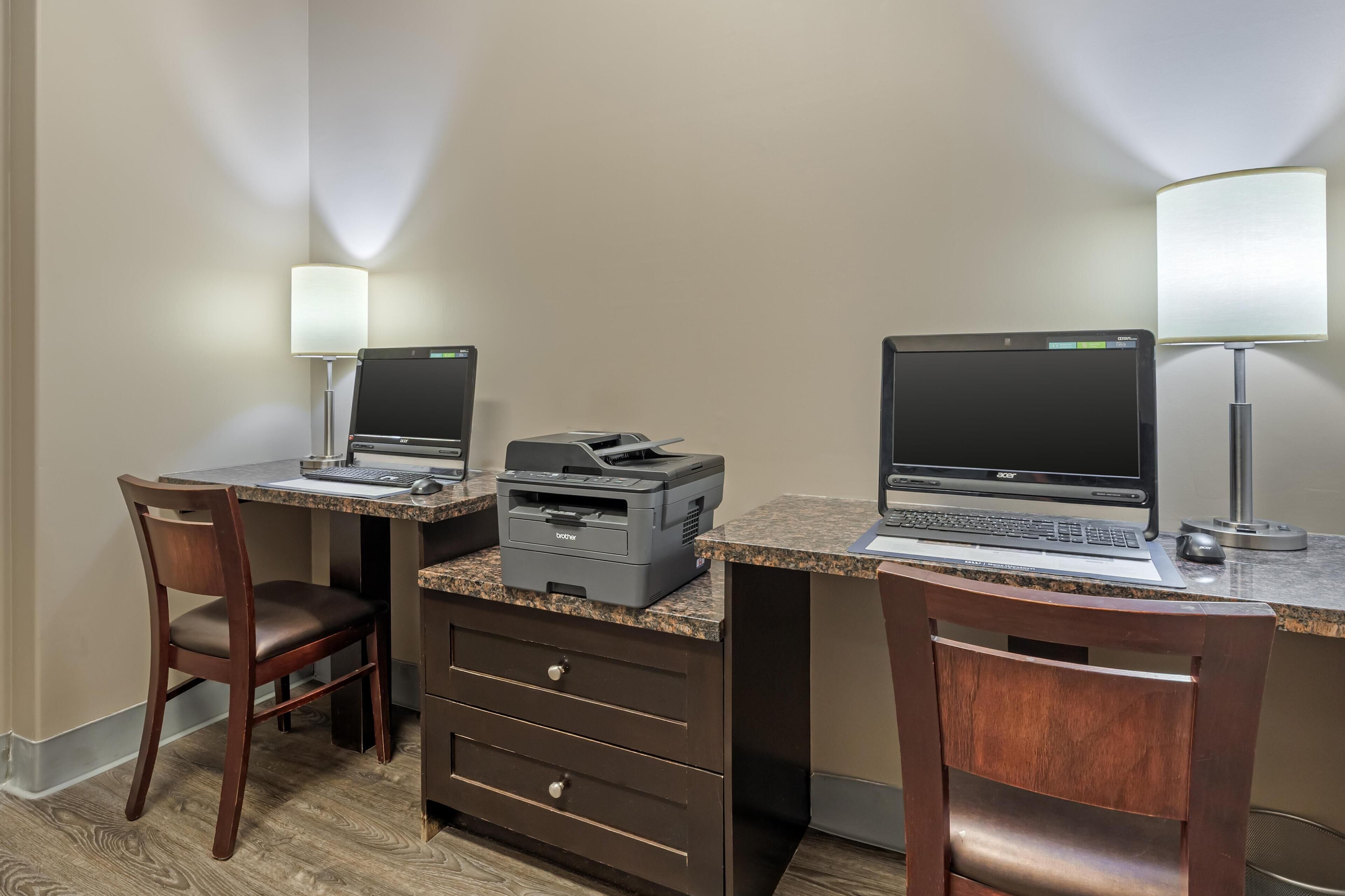 Een hotelkamer met twee bureaus, elk met een laptop en een printer, een stoel en een bureaulamp.