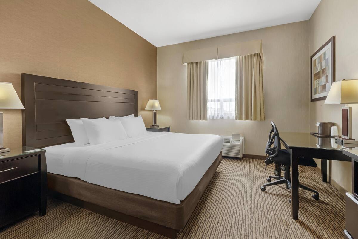 A hotel room with a large bed, bedside tables, a desk, a chair, a window with curtains, and a framed picture on the wall.