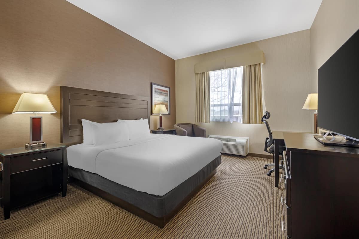 A hotel room with a large bed, two bedside tables with lamps, a desk with a chair, a TV, and a window with curtains.
