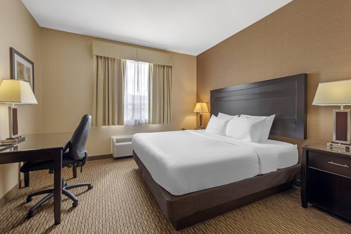 A hotel room with a large bed, a desk, two lamps, a chair, and a window with curtains.