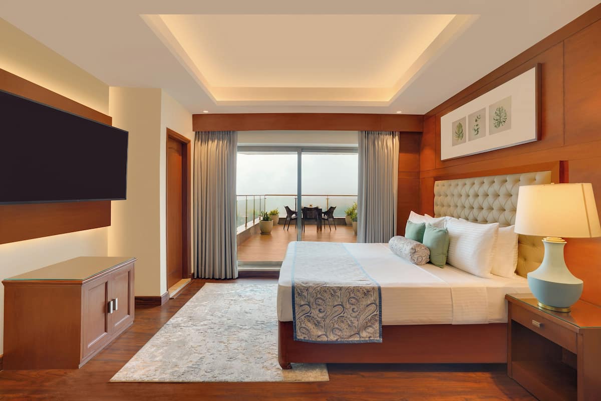 A hotel room with a large bed, a TV mounted on the wall, a bedside table with a lamp, and a view of the balcony with outdoor furniture.