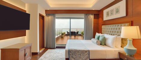 A hotel room with a large bed, a TV mounted on the wall, a bedside table with a lamp, and a view of the balcony with outdoor furniture.