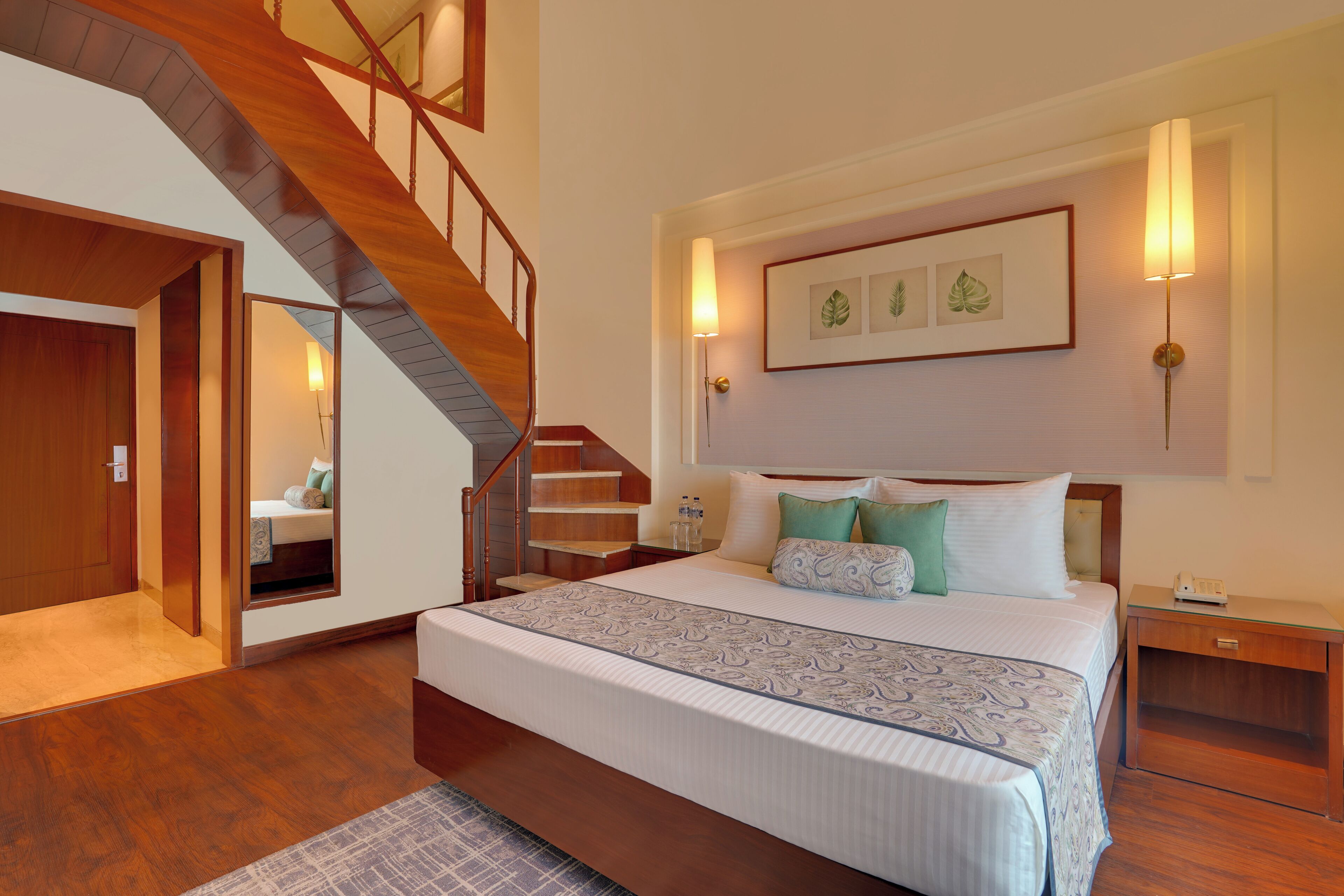 A bedroom with a large bed, a wooden staircase, a nightstand, and framed artwork on the wall.