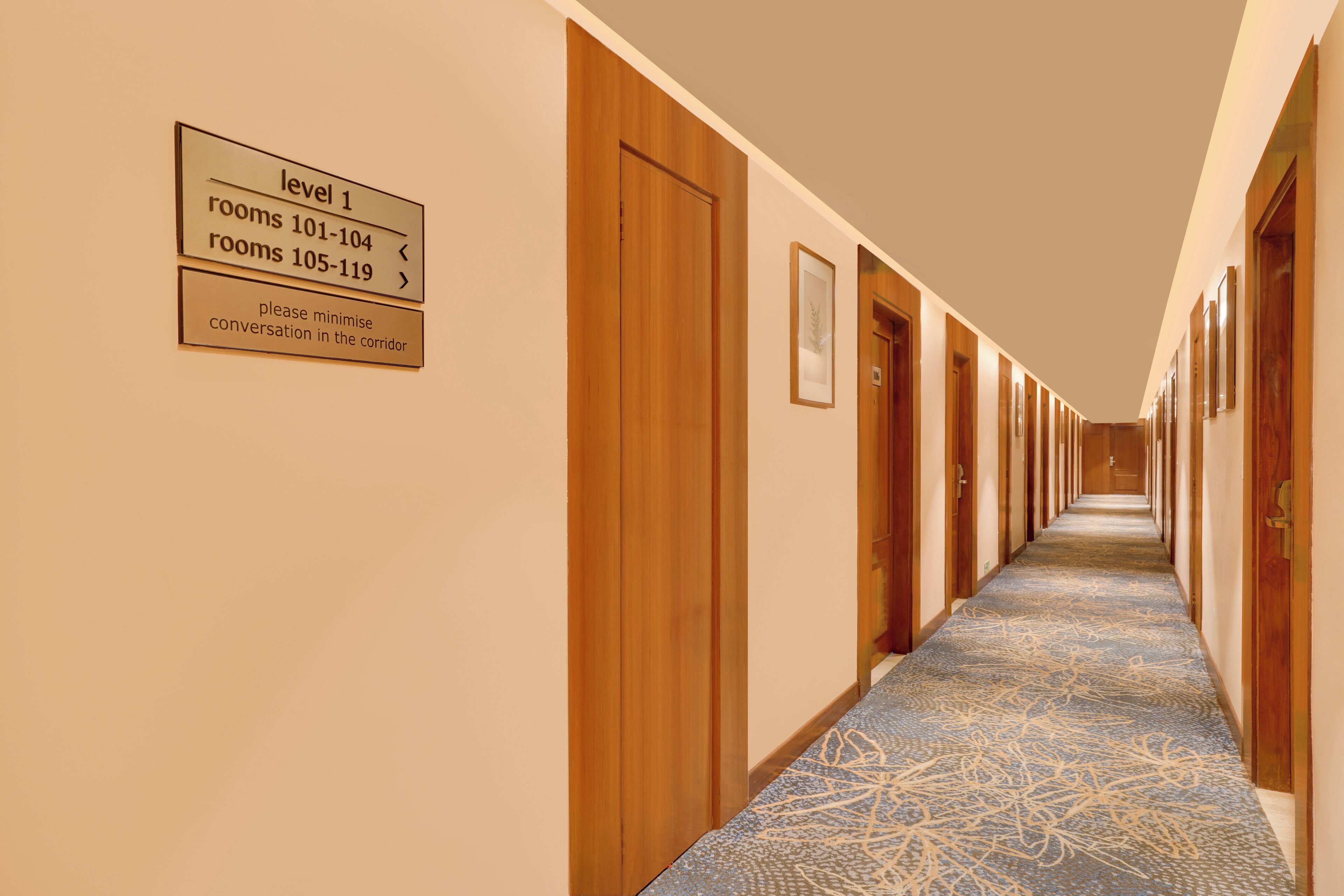A hotel corridor with wooden doors, a carpeted floor, and directional signs indicating room levels and numbers.