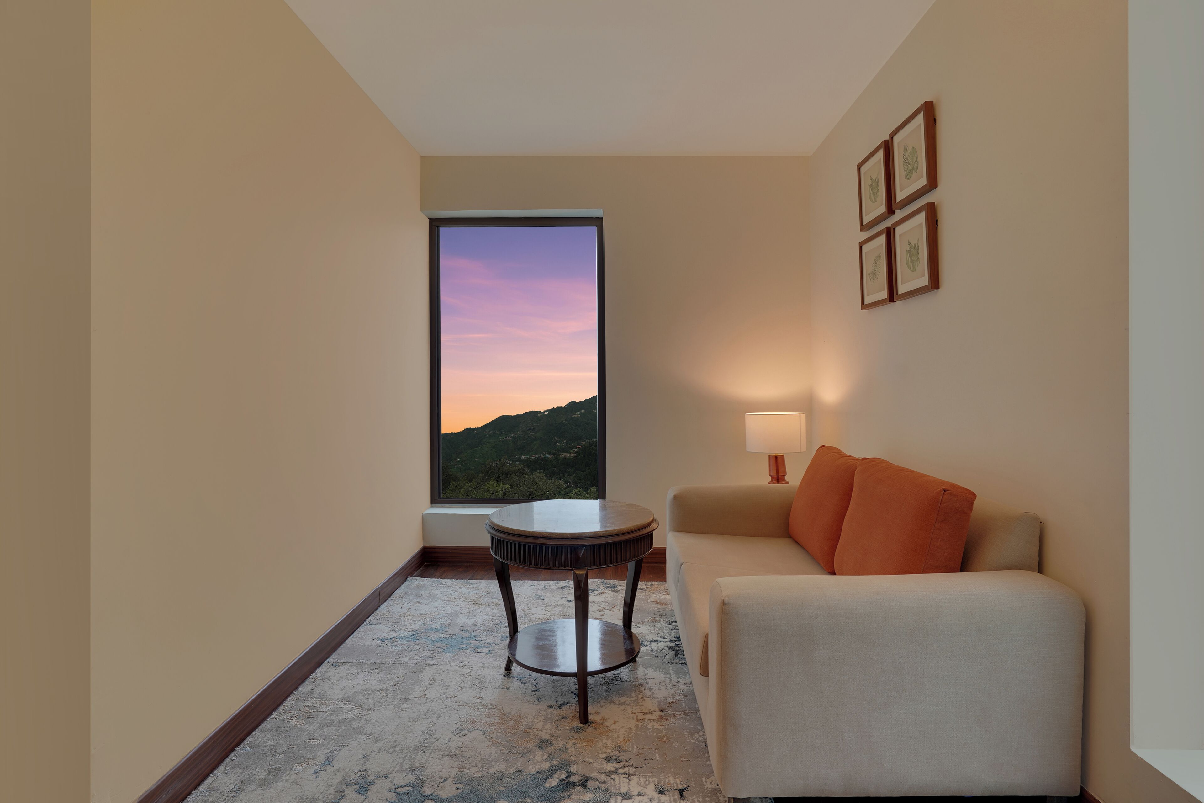 Ein modernes Hotelzimmer mit großem Fenster und Panoramablick auf einen Sonnenuntergang über Bergen, einem runden Holztisch, einem beigefarbenen Sofa mit orangefarbenen Kissen und einer Wandleuchte.
