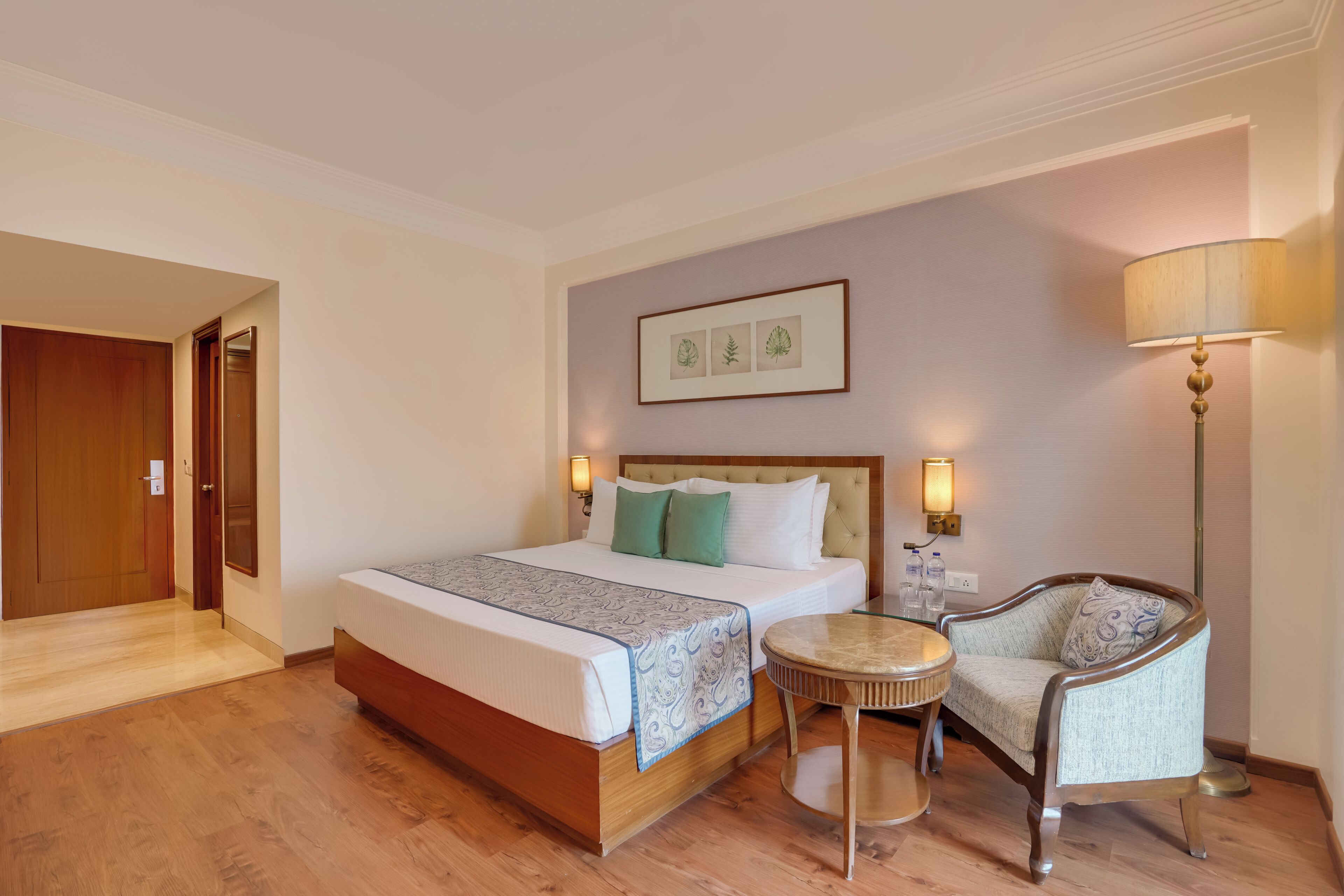 A hotel room with a bed, a chair, a small round table, and a wall-mounted lamp.