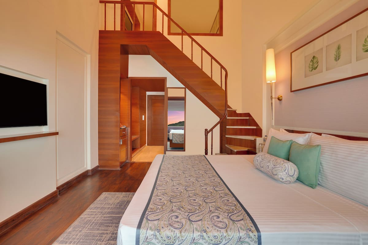A bedroom with a large bed, a wooden staircase, and a view of a sunset through an open door.
