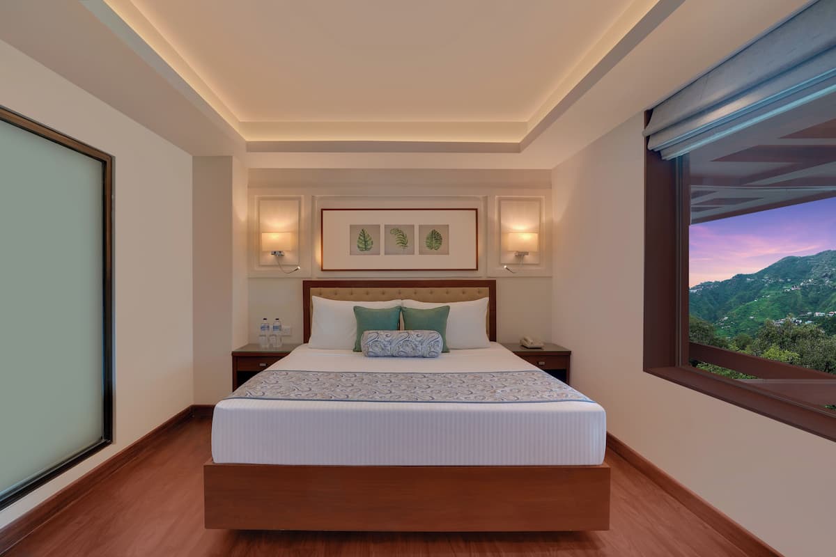 A modern hotel room with a large bed, bedside tables, a view of the mountains, and framed artwork on the wall.