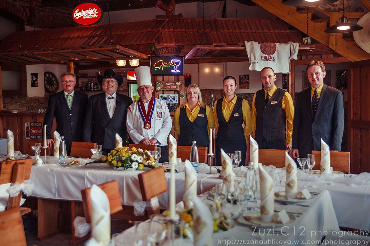 Eine Gruppe von Personen in einem Restaurant mit Holzvertäfelung, Tischen mit weißen Tischdecken und Kerzen, sowie einem Neonzeichen darüber.