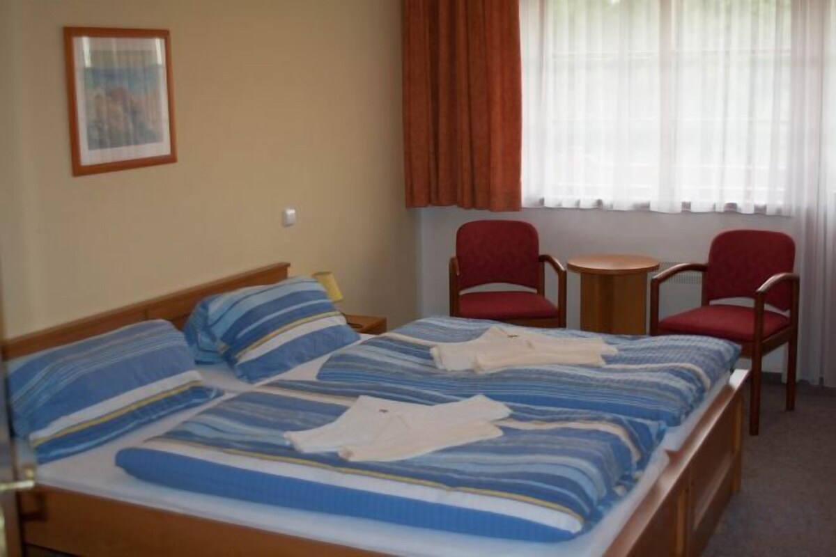 A double bed with striped bedding, a small round table, two red chairs, and a window with curtains.