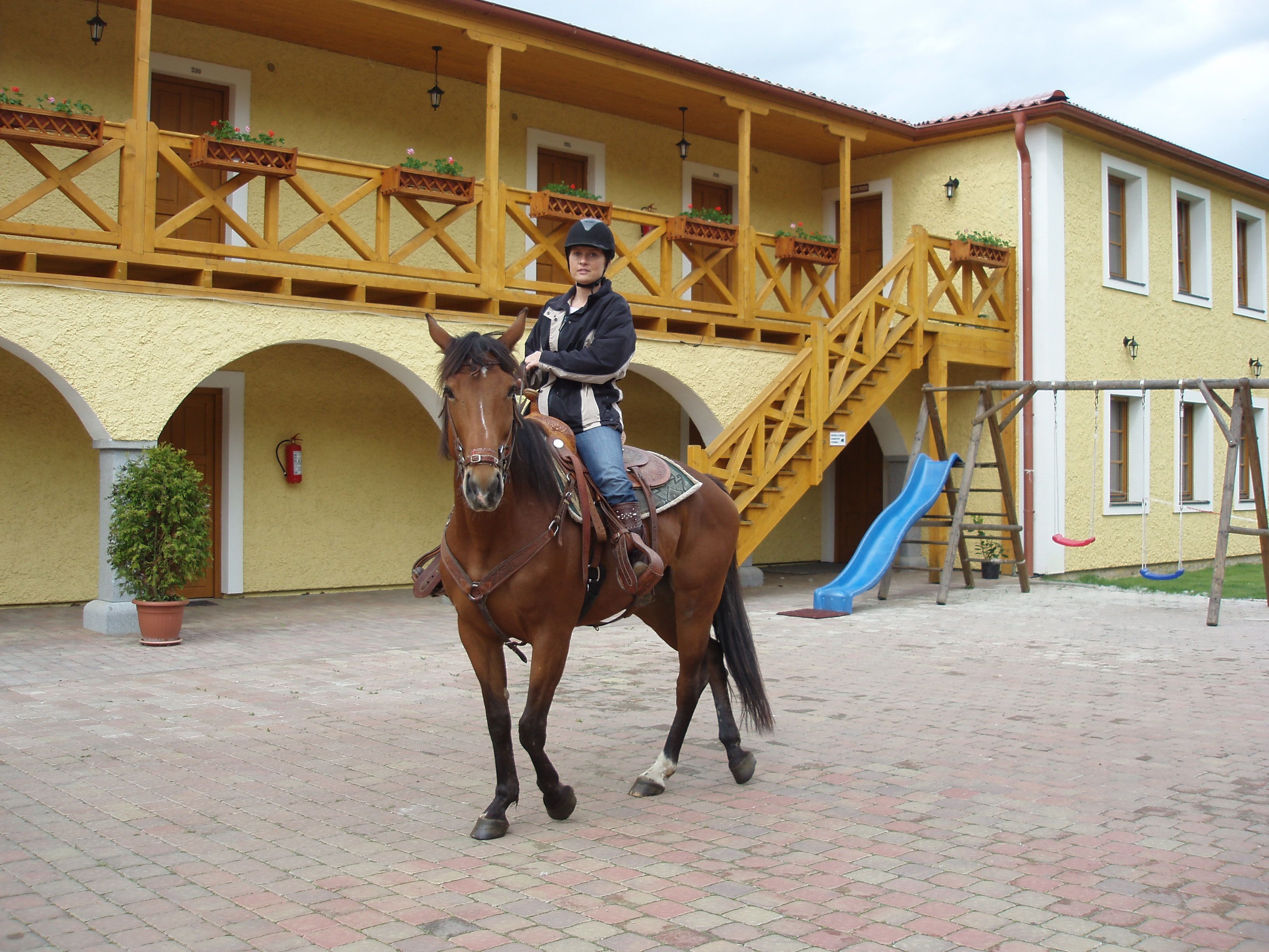 A horse and rider in an outdoor area with a yellow building, a wooden staircase, and a blue slide.