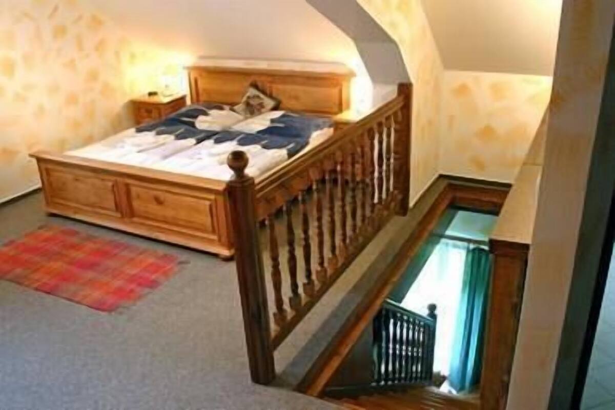 A bedroom with a wooden bed, a nightstand, a red plaid rug, and a staircase leading to an upper level.