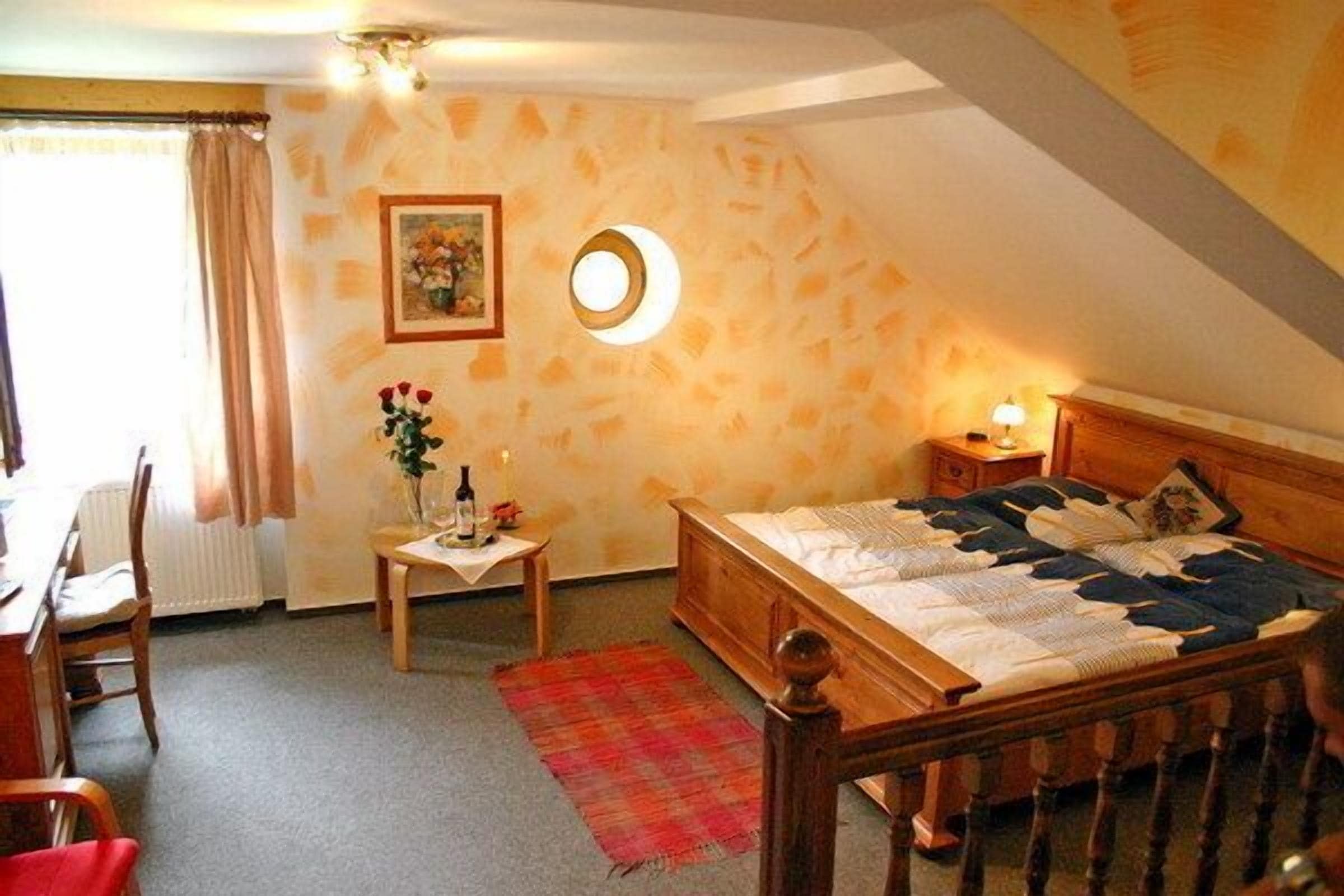 A bedroom with a wooden bed, a small bedside table, a nightstand, a red rug, a window with curtains, and a wall-mounted circular light fixture.