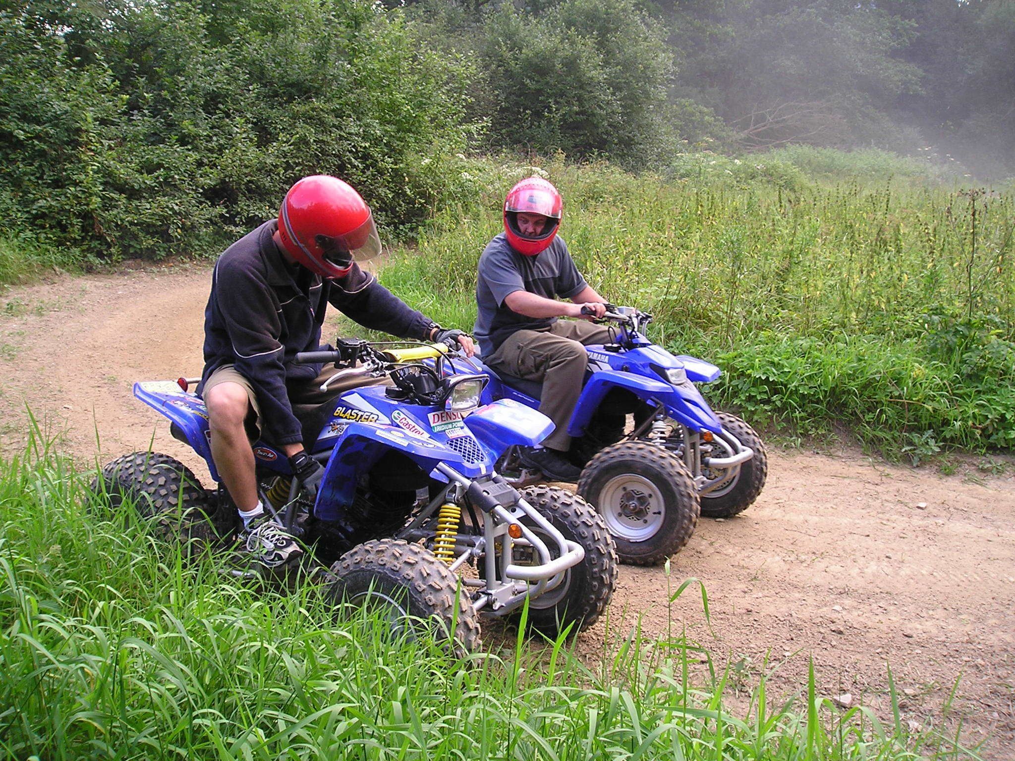 Two people on a blue ATV on a dirt path surrounded by greenery.
