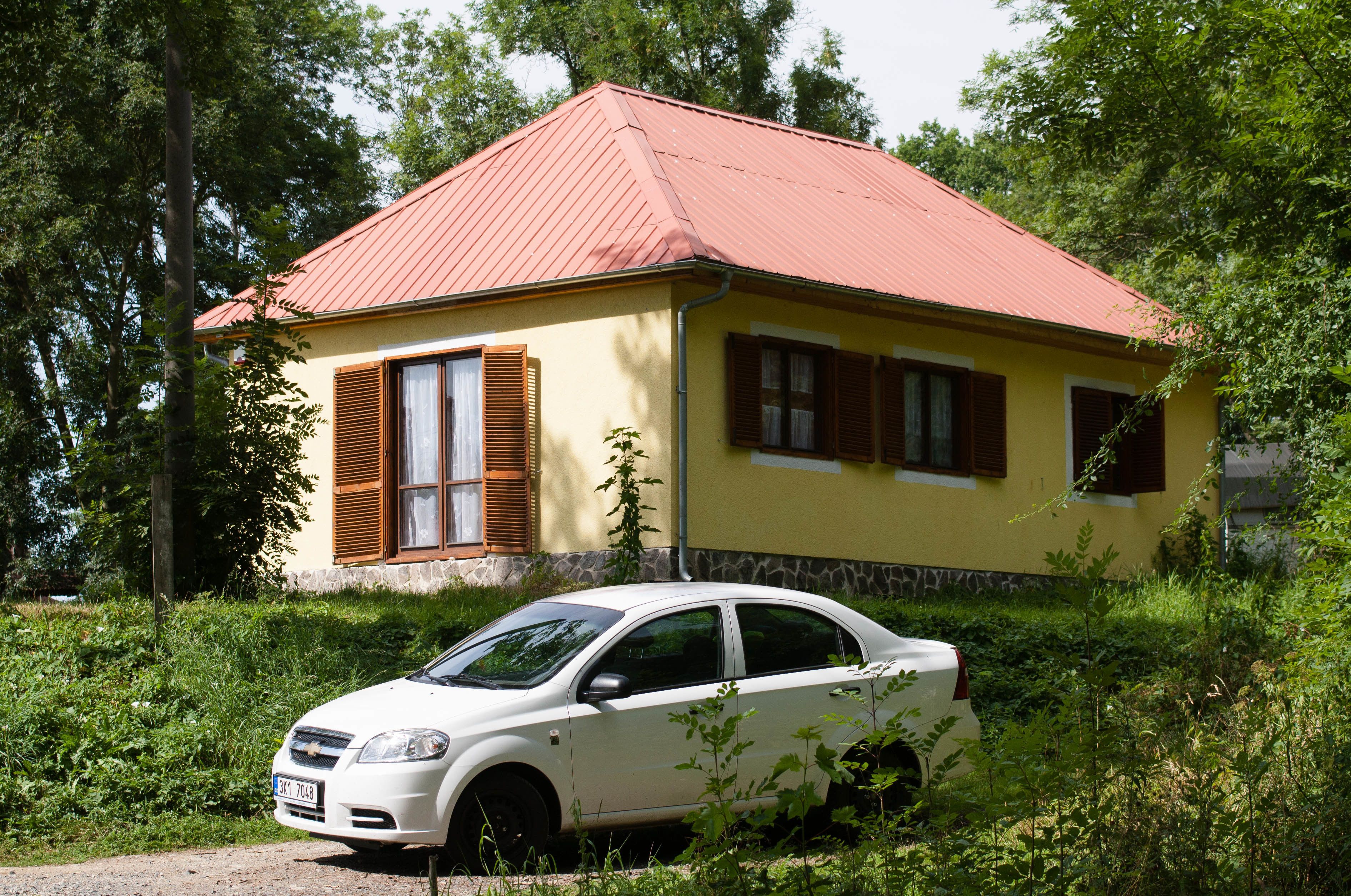 A yellow building with a red roof, a white car parked in front, and greenery around.