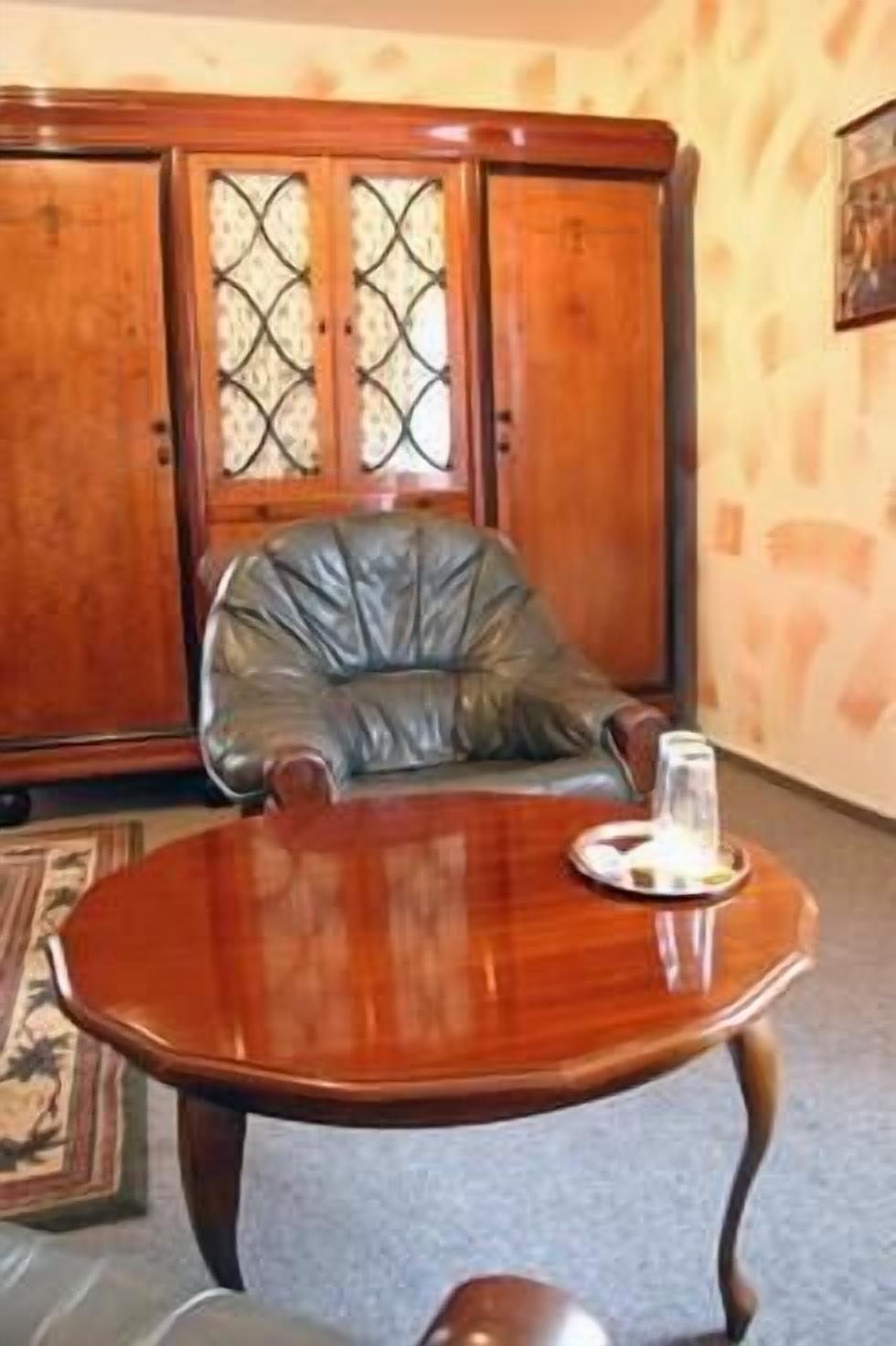 A wooden cabinet with glass doors, a leather chair, and a round wooden table with a glass on it.