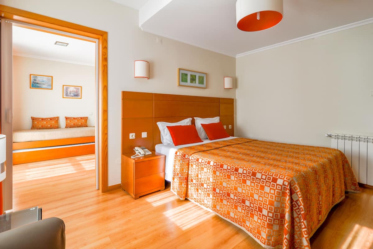 A hotel room with a wooden floor, a bed with an orange patterned bedspread, a nightstand with a phone, and a door leading to another room.