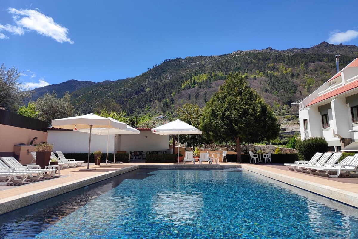 A swimming pool with lounge chairs and umbrellas, surrounded by a mountainous landscape.