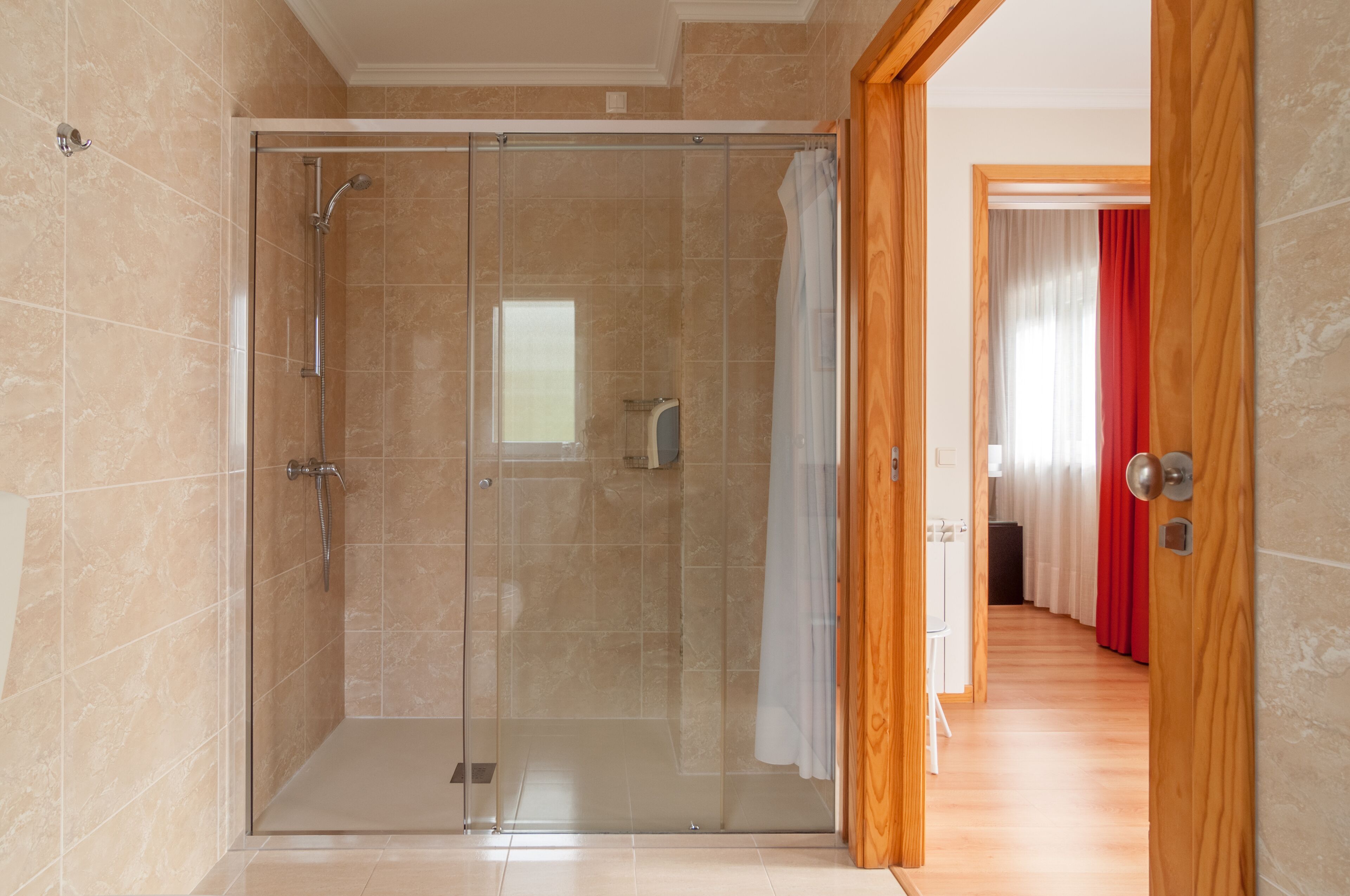 A glass-enclosed shower stall with a showerhead and control panel, adjacent to a wooden door leading to a hallway with a red curtain.