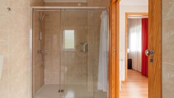 A glass-enclosed shower stall with a showerhead and control panel, adjacent to a wooden door leading to a hallway with a red curtain.